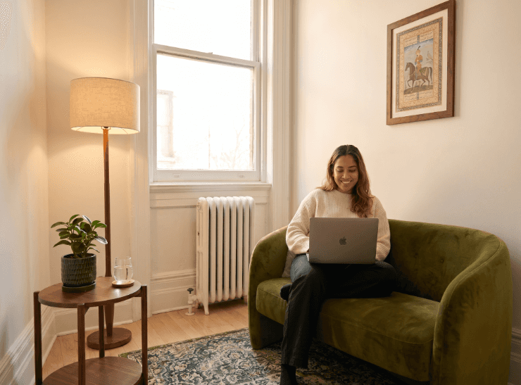 A woman relaxing on a green sofa in a sunlit room, using a laptop