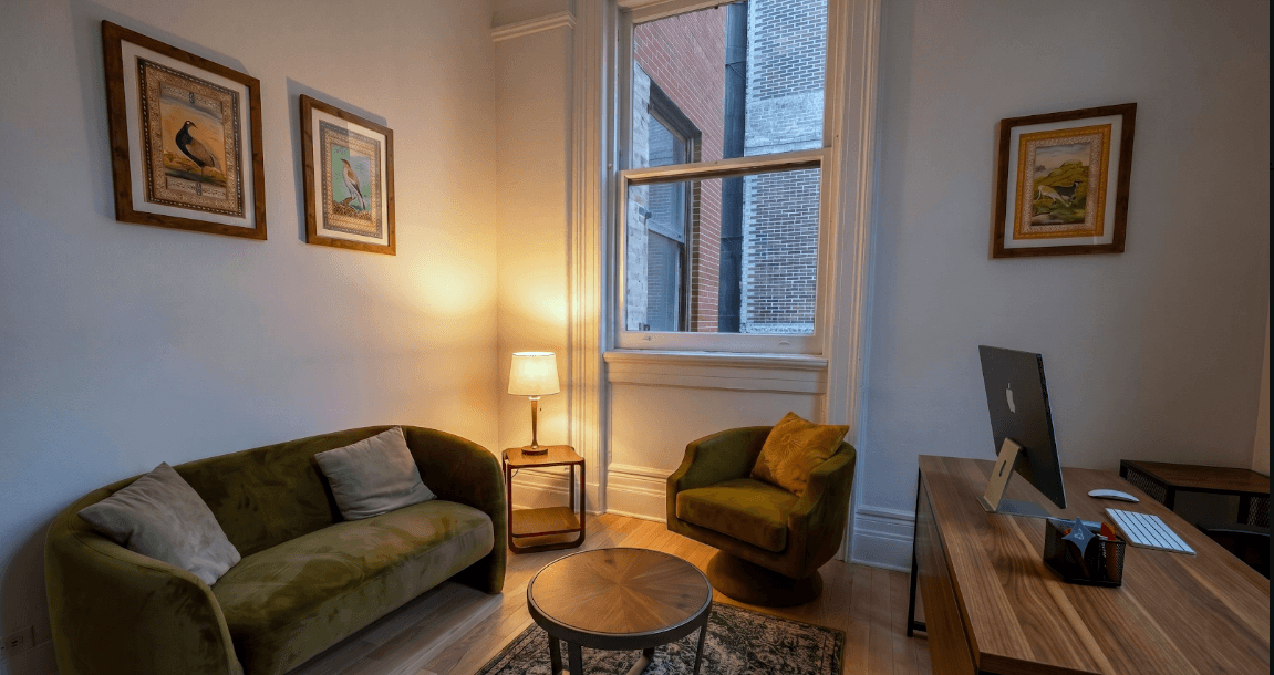 A cozy therapy waiting room with an olive-green sofa, a floor lamp, framed wall art and a bright window