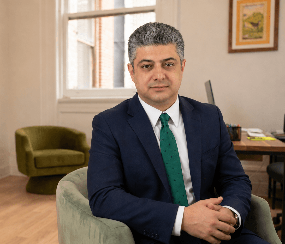 Dr. Omidvari in a navy suit and green tie, seated beside an olive armchair by a bright window