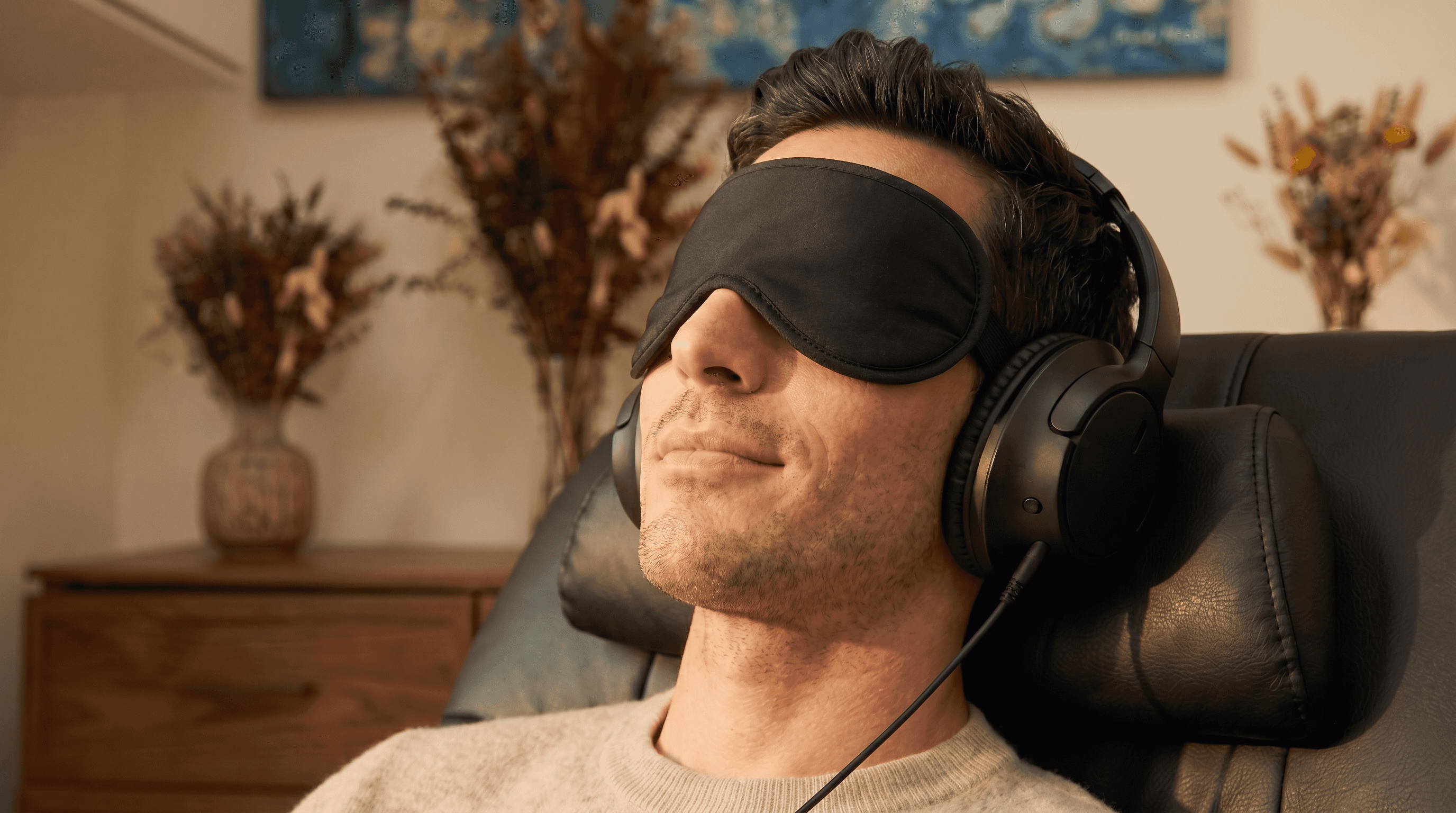 A man reclining in a leather chair wearing a black sleep eye-mask and over-ear headphones in a warm, dim room