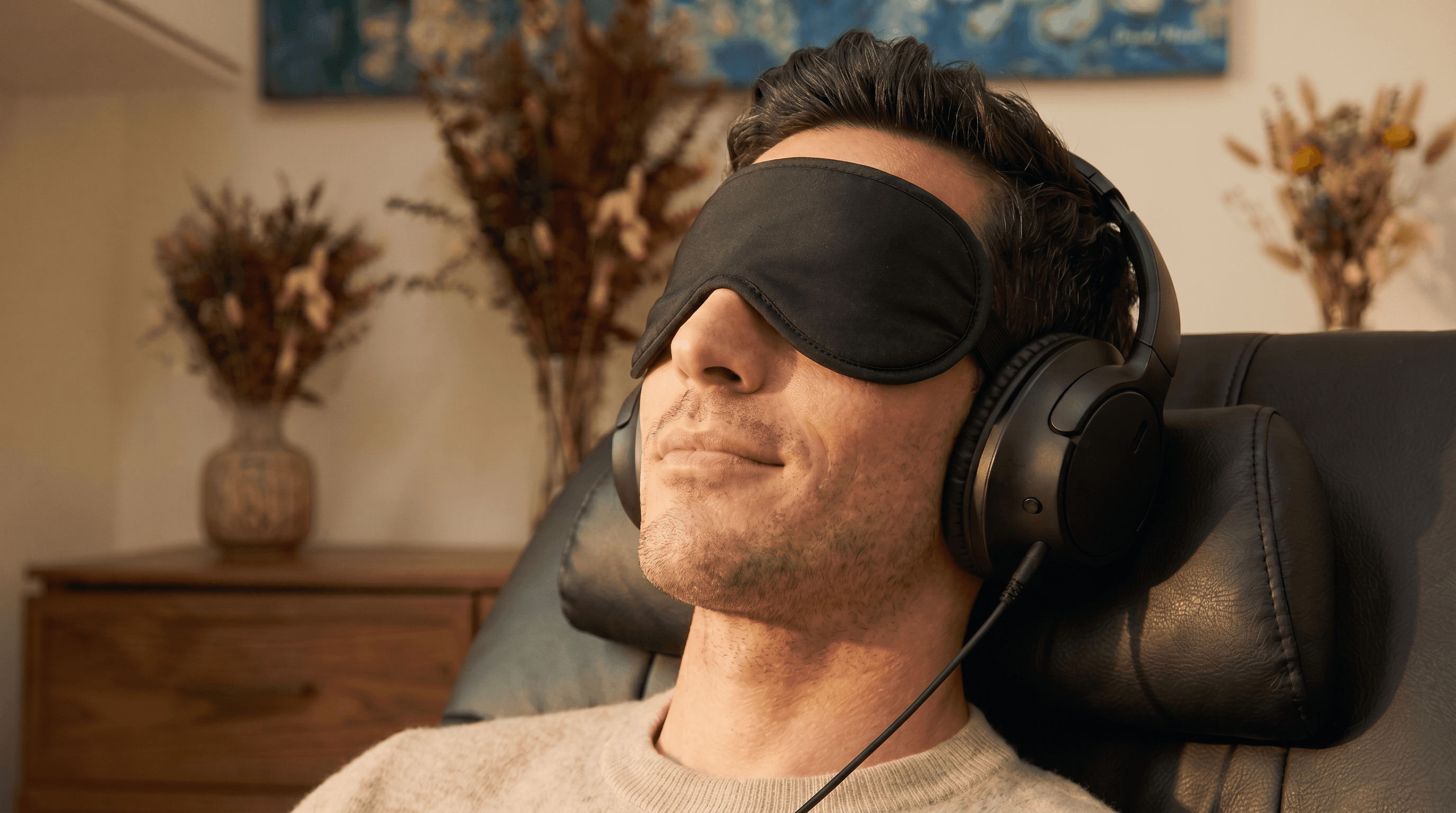 A man reclining in a leather chair wearing a black sleep eye-mask and over-ear headphones in a warm, dim room