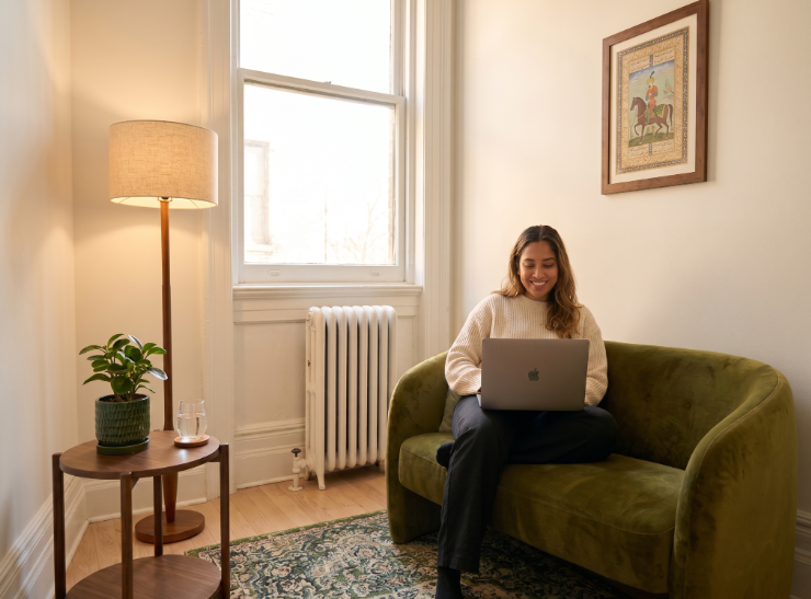 A woman relaxing on a green sofa in a sunlit room, using a laptop
